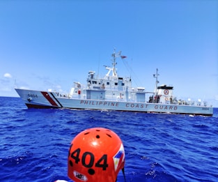 A Philippine Coast Guard vessel sails on a vibrant blue ocean. The ship is white with red stripes and the words 'PHILIPPINE COAST GUARD' are prominently displayed. A figure wearing an orange helmet labeled '4404' is visible in the foreground.