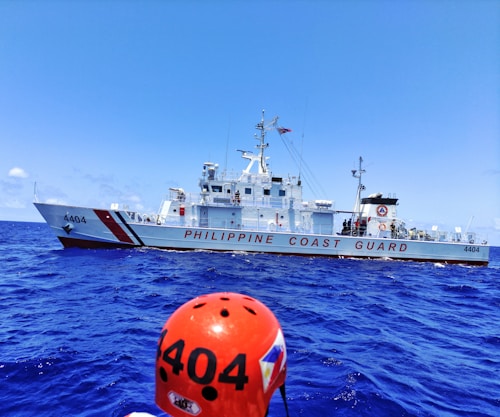 A Philippine Coast Guard vessel sails on a vibrant blue ocean. The ship is white with red stripes and the words 'PHILIPPINE COAST GUARD' are prominently displayed. A figure wearing an orange helmet labeled '4404' is visible in the foreground.
