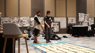 Two individuals stand in a room filled with large sheets of paper arranged on the floor. A group of people is seated on the carpeted floor, some engaging with the materials on the walls, which are covered in various posters and papers. The atmosphere suggests a workshop or collaborative event.