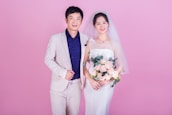 A couple is standing together, likely during a wedding. The woman is wearing a traditional white bridal gown with a veil and holding a bouquet of pink roses. The man is dressed in a light beige suit with a dark blue shirt. Both are smiling and positioned against a solid pink background.
