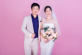 A couple is standing together, likely during a wedding. The woman is wearing a traditional white bridal gown with a veil and holding a bouquet of pink roses. The man is dressed in a light beige suit with a dark blue shirt. Both are smiling and positioned against a solid pink background.
