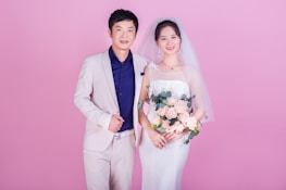 A couple is standing together, likely during a wedding. The woman is wearing a traditional white bridal gown with a veil and holding a bouquet of pink roses. The man is dressed in a light beige suit with a dark blue shirt. Both are smiling and positioned against a solid pink background.