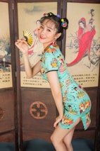 A woman dressed in a colorful, modern cheongsam with cartoon designs poses playfully while holding a gold fan in front of a traditional Asian-style folding screen featuring painted figures in historical attire.