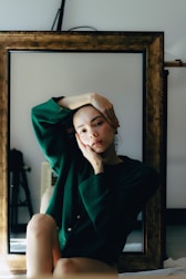 A person sits in front of a large rectangular mirror with a wooden frame, resting their head on one hand while the other arm is raised above their head. They are wearing a dark green button-up shirt and have a relaxed expression, creating a contemplative mood. The background is dimly lit with minimal visible objects, enhancing the focus on the person.