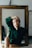 A person sits in front of a large rectangular mirror with a wooden frame, resting their head on one hand while the other arm is raised above their head. They are wearing a dark green button-up shirt and have a relaxed expression, creating a contemplative mood. The background is dimly lit with minimal visible objects, enhancing the focus on the person.