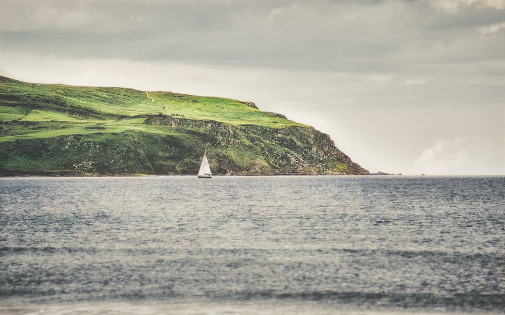 velero blanco en el mar cerca de la montaña verde bajo nubes blancas durante el día