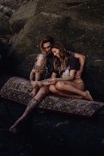 A couple sits on a patterned rug against a rocky backdrop, sharing an intimate moment while looking at a piece of paper. The man has tattoos and long hair, while the woman has long brown hair and is wearing casual attire. Both appear relaxed and content.