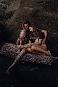 A couple sits on a patterned rug against a rocky backdrop, sharing an intimate moment while looking at a piece of paper. The man has tattoos and long hair, while the woman has long brown hair and is wearing casual attire. Both appear relaxed and content.