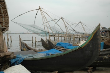 Several traditional fishing nets with large wooden frames are set up along a waterfront, with small wooden boats nearby. The scene appears to be in a fishing village with calm water and overcast skies.