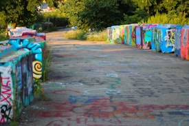 The image depicts a paved pathway flanked by low walls covered in colorful graffiti art. The path stretches into the distance, surrounded by vibrant green foliage and trees. The graffiti features various styles and colors, adding a lively and artistic feel to the scene.
