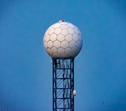 A tall radar tower features a spherical dome with a hexagonal pattern sitting atop a lattice metal structure against a clear blue sky.
