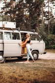 A person is standing beside an open door of a vintage camper van parked in a wooded area. The scene appears relaxed, indicating a possible camping or road trip scenario. The van is equipped for travel with a rooftop cargo box and features wooden elements inside.