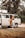 A person is standing beside an open door of a vintage camper van parked in a wooded area. The scene appears relaxed, indicating a possible camping or road trip scenario. The van is equipped for travel with a rooftop cargo box and features wooden elements inside.