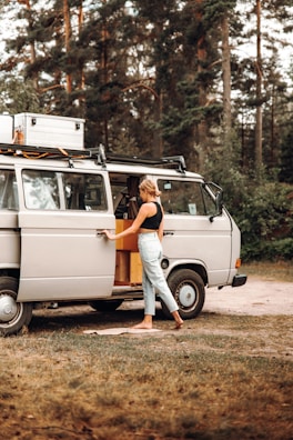 A person is standing beside an open door of a vintage camper van parked in a wooded area. The scene appears relaxed, indicating a possible camping or road trip scenario. The van is equipped for travel with a rooftop cargo box and features wooden elements inside.