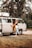 A person is standing beside an open door of a vintage camper van parked in a wooded area. The scene appears relaxed, indicating a possible camping or road trip scenario. The van is equipped for travel with a rooftop cargo box and features wooden elements inside.