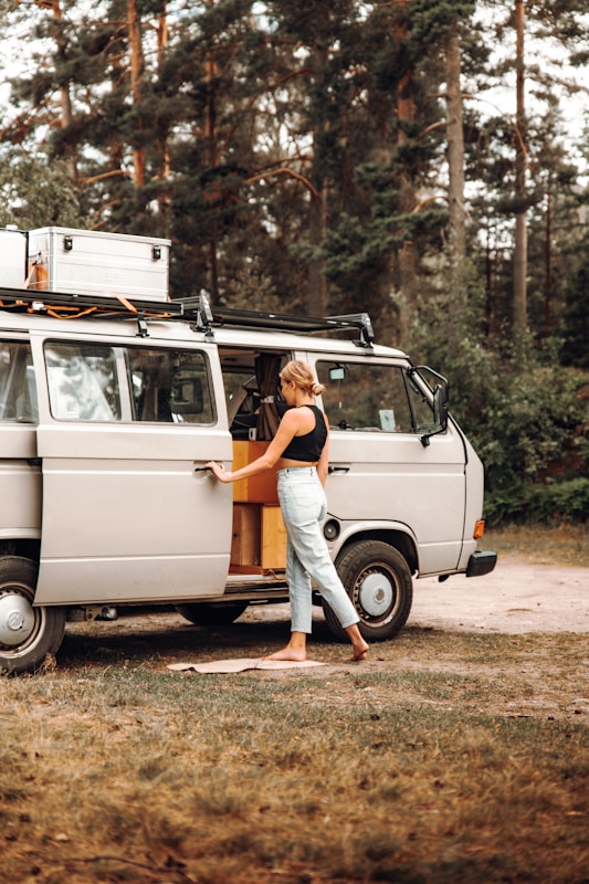 A person is standing beside an open door of a vintage camper van parked in a wooded area. The scene appears relaxed, indicating a possible camping or road trip scenario. The van is equipped for travel with a rooftop cargo box and features wooden elements inside.