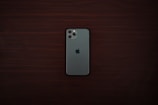 A smartphone with a greenish-gray back and a triple camera system is positioned centrally on a dark wooden surface. The smartphone bears a distinct logo of a partially eaten fruit in the center.