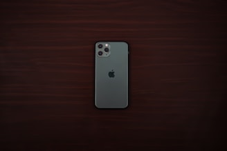 A smartphone with a greenish-gray back and a triple camera system is positioned centrally on a dark wooden surface. The smartphone bears a distinct logo of a partially eaten fruit in the center.