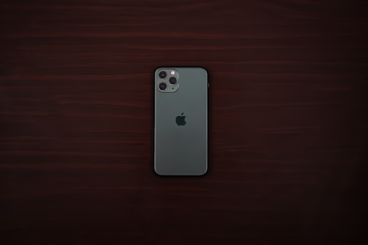 A smartphone with a greenish-gray back and a triple camera system is positioned centrally on a dark wooden surface. The smartphone bears a distinct logo of a partially eaten fruit in the center.