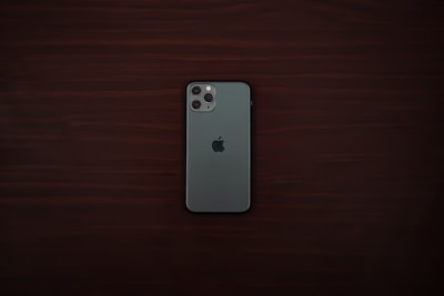 A smartphone with a greenish-gray back and a triple camera system is positioned centrally on a dark wooden surface. The smartphone bears a distinct logo of a partially eaten fruit in the center.