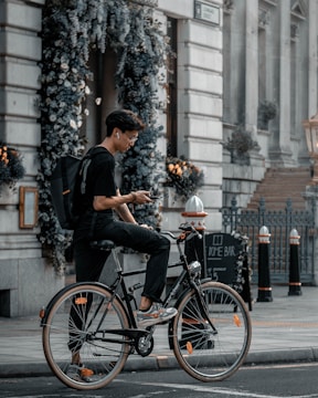 A person wearing wireless earbuds and casual clothing rides a bicycle on a city street. They are holding a smartphone, seemingly engaged in an activity, while a large backpack rests on their back. The background features a decorative archway adorned with flowers, a sidewalk, and the facade of a building with steps leading up. A signboard for a bar is visible, adding to the urban setting.