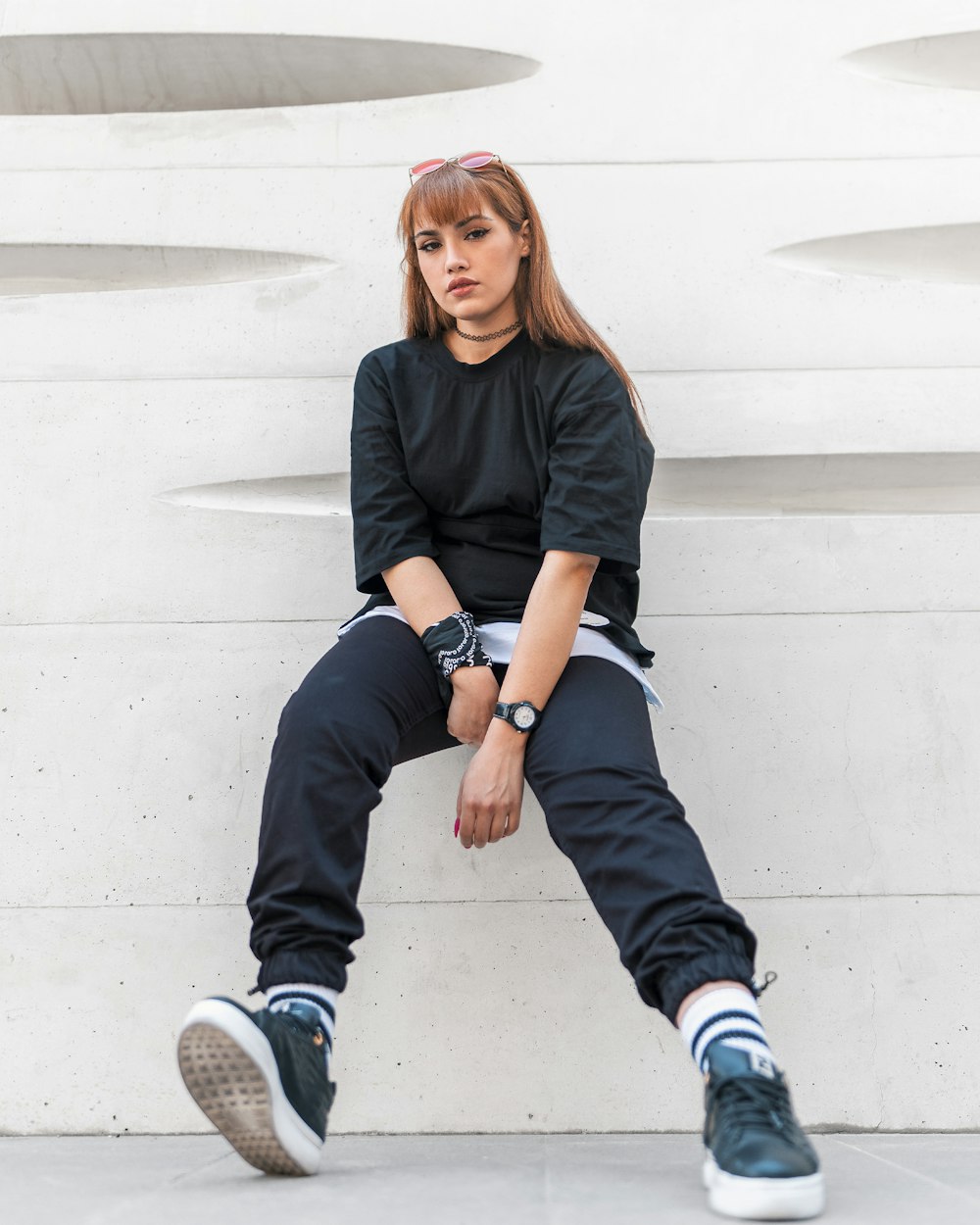 Mujer con camisa negra de manga larga y jeans de mezclilla azul sentada en una pared de concreto blanco