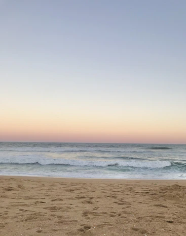 A wide-angle shot of a calm beach with gentle waves under a pastel sunset sky.