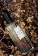 A perfume bottle labeled 'Gift of Plant Eau de Parfum Sunit Ricefield' is placed among dried flowers and plants, creating a rustic and natural setting. The bottle is made of clear glass with a black cap, and the label is simple and elegant.