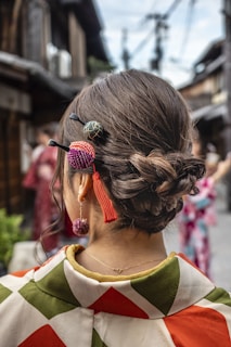A woman wearing a traditional kimono with an intricate bun hairstyle adorned with colorful hair ornaments. The background features a blurred view of a street, with traditional architecture and another person in a kimono.