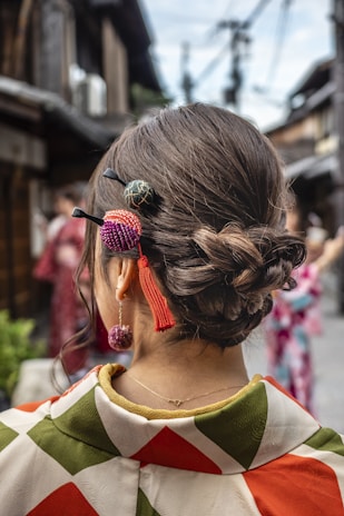 A woman wearing a traditional kimono with an intricate bun hairstyle adorned with colorful hair ornaments. The background features a blurred view of a street, with traditional architecture and another person in a kimono.