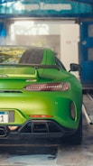 A vibrant green sports car is parked in a car wash area. The car has a sleek, aerodynamic design with a prominent rear spoiler and dual exhaust pipes. Water droplets are visible on the body of the car, indicating it has been recently washed. The surroundings show a blue overhang with signage.