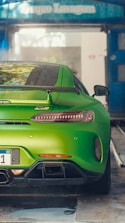A vibrant green sports car is parked in a car wash area. The car has a sleek, aerodynamic design with a prominent rear spoiler and dual exhaust pipes. Water droplets are visible on the body of the car, indicating it has been recently washed. The surroundings show a blue overhang with signage.