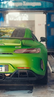 A vibrant green sports car is parked in a car wash area. The car has a sleek, aerodynamic design with a prominent rear spoiler and dual exhaust pipes. Water droplets are visible on the body of the car, indicating it has been recently washed. The surroundings show a blue overhang with signage.