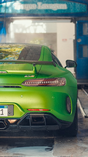 A vibrant green sports car is parked in a car wash area. The car has a sleek, aerodynamic design with a prominent rear spoiler and dual exhaust pipes. Water droplets are visible on the body of the car, indicating it has been recently washed. The surroundings show a blue overhang with signage.