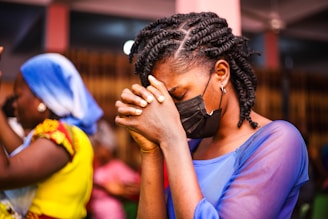 A person with braided hair, wearing a black face mask, is seen in a moment of prayer or contemplation with hands clasped together. The setting appears to be indoors, with other individuals in the background. One person is wearing a vibrant yellow outfit with a blue headscarf.