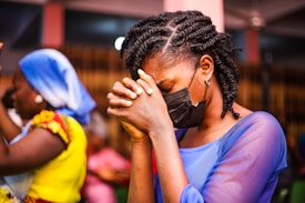 A person with braided hair, wearing a black face mask, is seen in a moment of prayer or contemplation with hands clasped together. The setting appears to be indoors, with other individuals in the background. One person is wearing a vibrant yellow outfit with a blue headscarf.