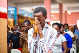 A group of people gathered indoors, engaged in an activity that seems to involve contemplation or prayer. The central figure is a man in a striped shirt, with hands clasped together in front of his face and eyes closed. Other individuals in the background appear to be in similar postures, suggesting a unified or communal experience.