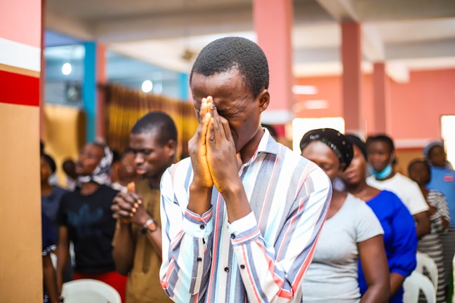 A group of people gathered indoors, engaged in an activity that seems to involve contemplation or prayer. The central figure is a man in a striped shirt, with hands clasped together in front of his face and eyes closed. Other individuals in the background appear to be in similar postures, suggesting a unified or communal experience.