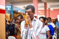 A group of people gathered indoors, engaged in an activity that seems to involve contemplation or prayer. The central figure is a man in a striped shirt, with hands clasped together in front of his face and eyes closed. Other individuals in the background appear to be in similar postures, suggesting a unified or communal experience.