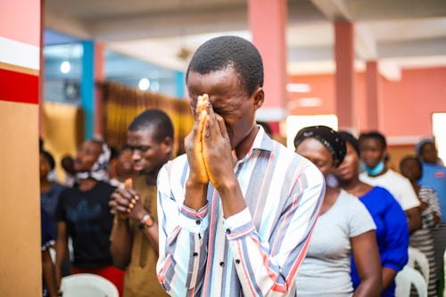 A group of people gathered indoors, engaged in an activity that seems to involve contemplation or prayer. The central figure is a man in a striped shirt, with hands clasped together in front of his face and eyes closed. Other individuals in the background appear to be in similar postures, suggesting a unified or communal experience.