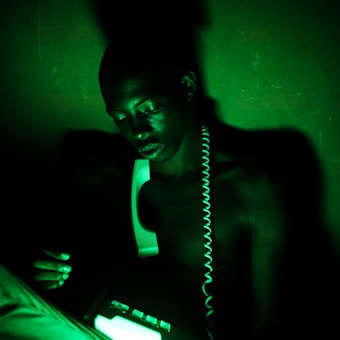 A person sits in a dimly lit setting, illuminated by a green light that casts dramatic shadows. They hold a telephone receiver with the coiled cord draped around their neck, creating a mysterious atmosphere.