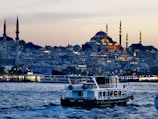 A scenic view of a bustling waterfront in the evening, featuring a boat with a Turkish flag prominently displayed in the foreground. The background showcases an illuminated cityscape with grand mosques and minarets, highlighting Islamic architecture against a dusky sky.