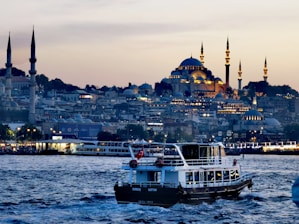 A scenic view of a bustling waterfront in the evening, featuring a boat with a Turkish flag prominently displayed in the foreground. The background showcases an illuminated cityscape with grand mosques and minarets, highlighting Islamic architecture against a dusky sky.