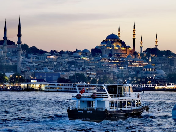 A scenic view of a bustling waterfront in the evening, featuring a boat with a Turkish flag prominently displayed in the foreground. The background showcases an illuminated cityscape with grand mosques and minarets, highlighting Islamic architecture against a dusky sky.