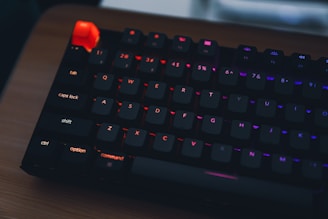 A mechanical keyboard with RGB backlighting features prominently lit keys against a dark background. The keys emit a range of colors including red, blue, and orange. The keyboard is placed on a wooden surface.