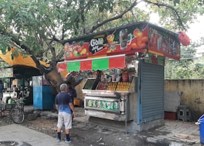 A street-side juice stand with colorful signage and a canopy selling various fruit juices and beverages. The stall displays an assortment of fresh fruits, including a pile of oranges and bottles of soft drinks. A man wearing shorts and a blue shirt is interacting with the vendor. Trees and a yellow canopy are in the background, with a bicycle parked nearby.