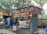 A street-side juice stand with colorful signage and a canopy selling various fruit juices and beverages. The stall displays an assortment of fresh fruits, including a pile of oranges and bottles of soft drinks. A man wearing shorts and a blue shirt is interacting with the vendor. Trees and a yellow canopy are in the background, with a bicycle parked nearby.