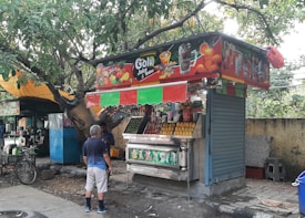A street-side juice stand with colorful signage and a canopy selling various fruit juices and beverages. The stall displays an assortment of fresh fruits, including a pile of oranges and bottles of soft drinks. A man wearing shorts and a blue shirt is interacting with the vendor. Trees and a yellow canopy are in the background, with a bicycle parked nearby.