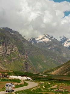 A scenic mountainous landscape features lush green valleys and snow-capped peaks beneath a sky filled with thick clouds. A colorful truck navigating a winding road adds a touch of human activity, and scattered tents hint at a temporary settlement against the rugged backdrop.
