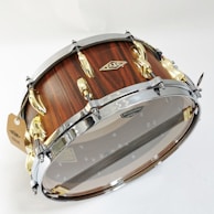 A beautifully crafted snare drum with a wooden finish and metal hardware. The shell displays a rich wood grain texture, complemented by polished chrome and gold hardware, including lugs and hoops. A transparent drumhead allows visibility of the internal components, such as the snare wires.